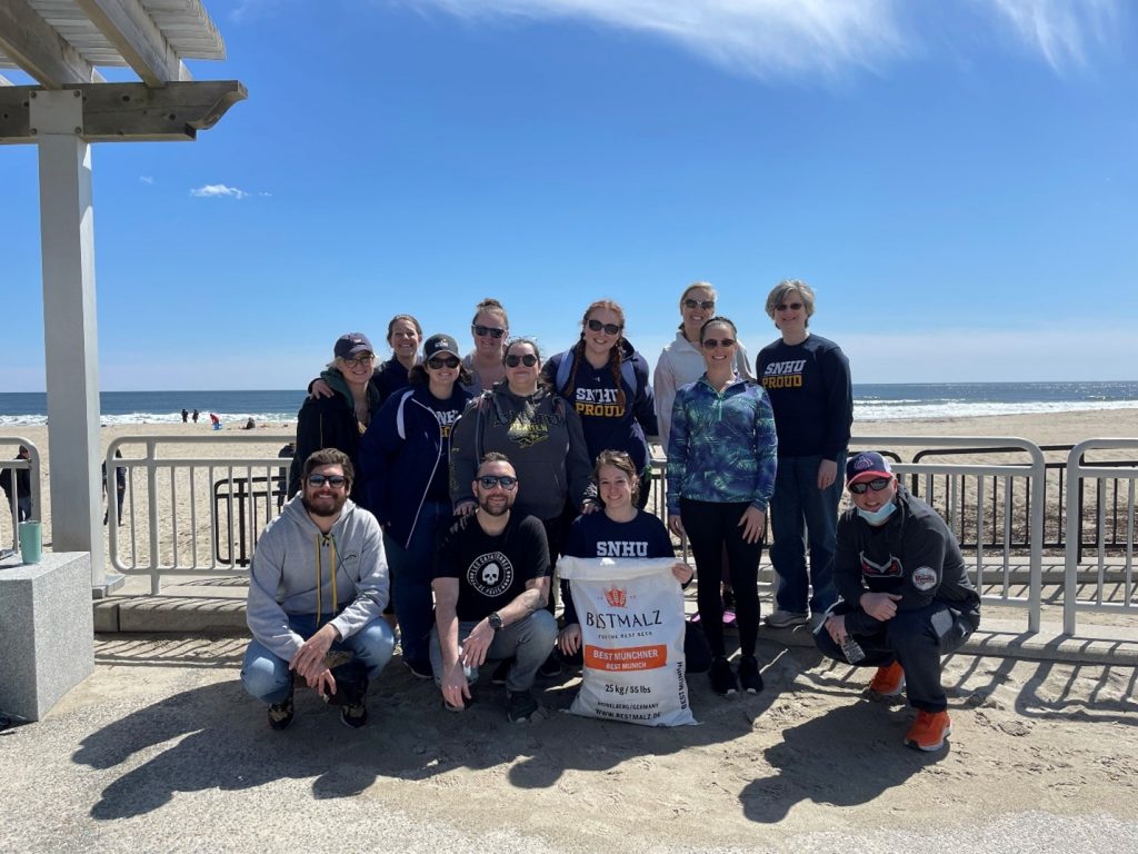 SNHU Academic Advisors after a cleanup at Hampton Beach. Shows a group of volunteers with a bag of trash, posed near the railing of the boardwalk at Hampton Beach.