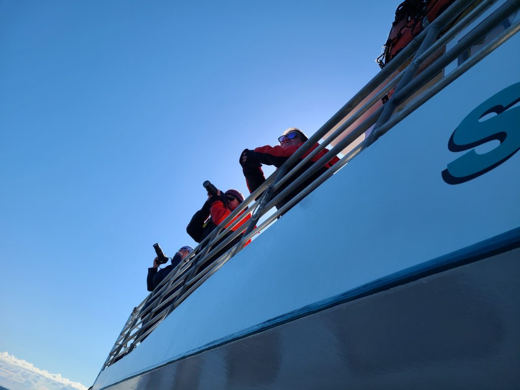 Scientists watching for whales from a survey cruise. Image shows 3 women scientists with cameras, looking over the side of a boat.