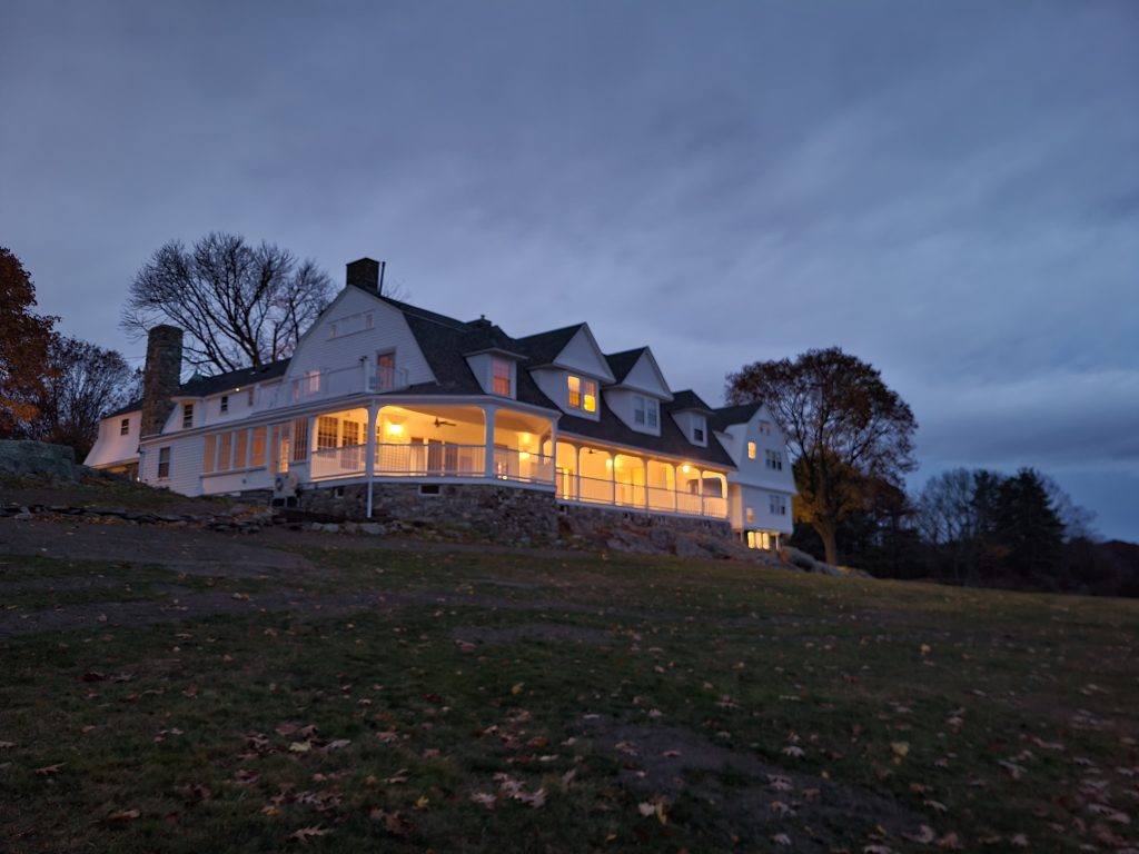 Carey Cottage, a large white building, shown from the water. It is dusk and the building is aglow with light. There is green lawn in the foreground. 